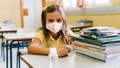 girl_sitting_at_her_chair_and_table_in_the_classroom_wearing_a_mask_to_protect_herself_during_the...