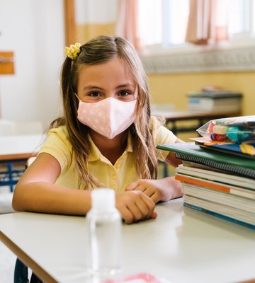 girl_sitting_at_her_chair_and_table_in_the_classroom_wearing_a_mask_to_protect_herself_during_the...