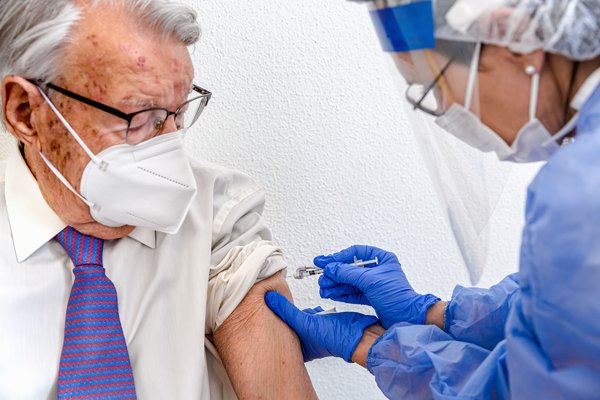 elderly_man_in_mask_watches_as_nurse_in_protective_suit_and_covid_mask_gives_him_coronavirus_vaccine