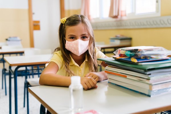 girl_sitting_at_her_chair_and_table_in_the_classroom_wearing_a_mask_to_protect_herself_during_the...